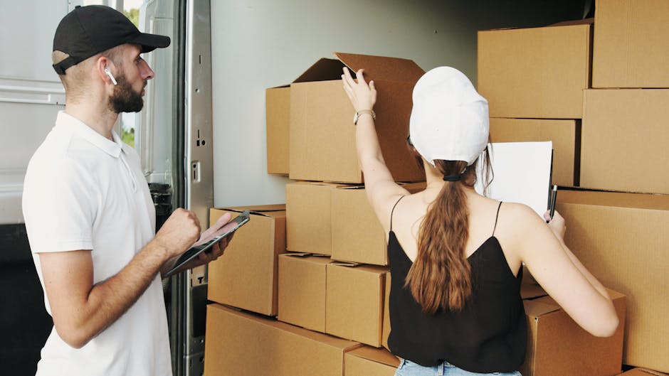A man and a woman are inside a vehicle, preparing for a home relocation with furniture and packed boxes around them. The man, wearing a white t-shirt, black cap, and earbuds, holds a clipboard and is observing the loading process. The woman, dressed in a black sleeveless top and white cap, is using a smartphone and appears to be inspecting or organizing the cardboard boxes stacked neatly inside the van. The boxes are of various sizes, made of cardboard, and some are partially open revealing packing materials like paper or plastic wrap. The scene takes place near the entrance of the vehicle, with the van's interior visible in the background, highlighting the packing and loading activities associated with furniture transport and house removals. Man with Van Coombe's professional services involve packing and moving, emphasizing careful handling of household items during the loading process in a professional move.