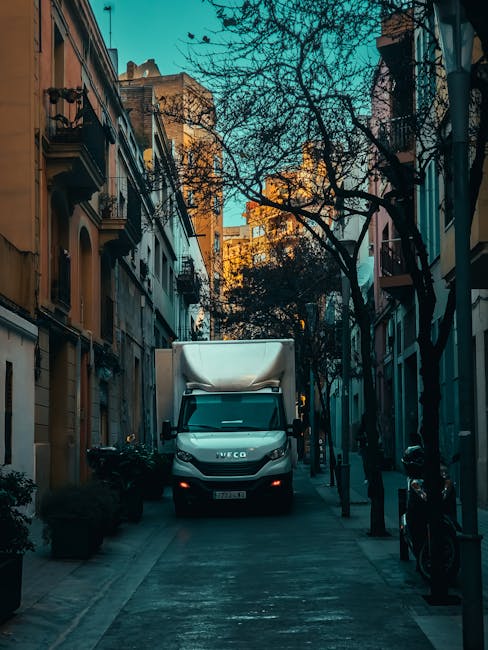 A black moving truck with its rear door open is parked on a narrow street in a residential area, partially on the pavement and partially on the road. The truck is in the process of being loaded, with some furniture and boxes visible inside. The street is lined with multi-storey houses featuring traditional architecture, with hanging flower baskets and a street lamp attached to a building on the right. A man is visible in the distance, standing near the truck, likely coordinating the move. Nearby, there is a small wooden bench with potted plants and decorative items placed on the sidewalk. The scene is overcast, suggesting typical UK weather, and the overall setting indicates a house removal or home relocation involving furniture transport and packing. Man with Van Coombe's services are implied through the context, supporting efficient moving logistics.