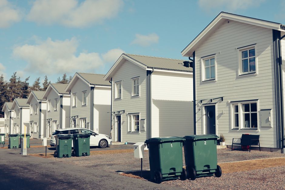 A row of modern, white, two-story residential houses with gabled roofs and multiple windows, situated in a suburban neighbourhood on a clear day with blue skies and scattered clouds. In the foreground, several dark green wheeled bins are positioned on the gravel driveway, each with labels indicating collection days, alongside a white mailbox. A white car is parked near the houses, partially visible, while a black moving blanket covers an item being carried by movers, and a hand truck or trolley is visible on the left side of the image. The scene depicts a typical home relocation setting, with the moving process involving the loading or unloading of household items. The environment appears clean and organized, with some benches near the houses, and the overall setting emphasizes house removals, furniture transport, and packing and moving activities as part of a residential move. Man with Van Coombe occasionally mentioned in context to the moving process.