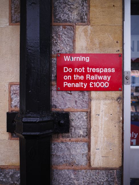 A close-up view of a brick and stone exterior wall next to a door frame. Mounted on the wall is a red warning sign with white text stating, 'Warning Do not trespass on the Railway Penalty £1000.' Adjacent to the sign is a black metal pipe running vertically along the wall, secured with brackets. This setting appears to be outside a building, possibly near a residential or commercial property, with the sign indicating railway trespass prohibitions. The image is relevant to house removals and relocating services, highlighting safety and legal considerations during home relocation or furniture transport processes managed by Man with Van Coombe.