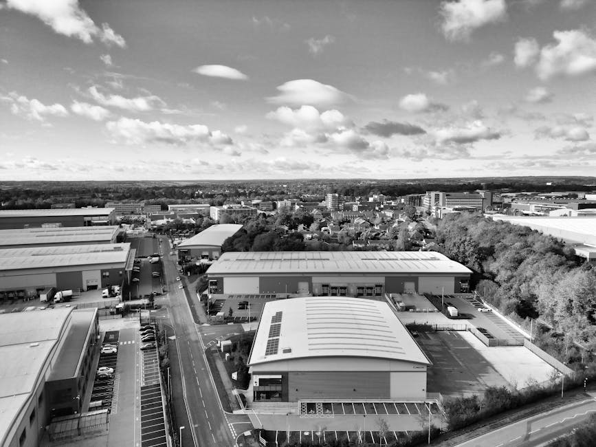 An aerial black-and-white photograph showcases a commercial or industrial area with multiple large warehouse-style buildings featuring flat roofs, some with solar panels, and surrounded by parking lot