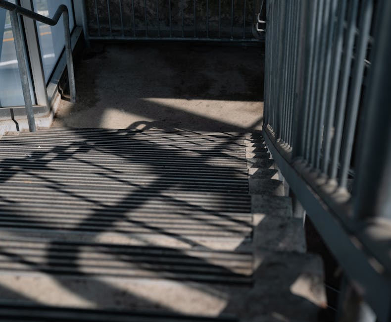 A metal staircase with grated steps leading down to an outdoor area, with cast shadow lines created by the railing. On the right, a tall metal railing runs parallel to the stairs, providing safety and support. The staircase is attached to a building or residential complex, indicated by the shadowed entrance at the top. The surrounding environment appears to be an external access point, possibly at the entrance or exit of a communal building or apartment block, relevant to house removals or moving logistics. The image showcases the structural details of the staircase and railings, important for assessing access and safety during a home relocation, with residual daylight highlighting the material and design. Man with Van Coombe may utilize such features when planning furniture transport and loading procedures, ensuring careful navigation of stairs during moves.