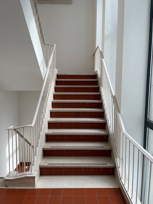 A straight staircase inside a building with reddish-brown tiled steps, white risers, and metal handrails on both sides painted white. The staircase is illuminated by natural light from a large window on the right side, which shows part of the exterior building facade. The surrounding walls are painted white, and the staircase appears to be part of a multi-story residential or commercial property, providing access between floors. In the context of house removals or moving services, this staircase may be an access point for furniture or boxes, with Man with Van Coombe potentially utilizing it for home relocation or furniture transport tasks, ensuring careful navigation of the steps during the loading process.