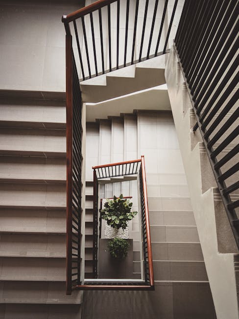 A metal staircase with grated steps leading down to an outdoor area, with cast shadow lines created by the railing. On the right, a tall metal railing runs parallel to the stairs, providing safety and support. The staircase is attached to a building or residential complex, indicated by the shadowed entrance at the top. The surrounding environment appears to be an external access point, possibly at the entrance or exit of a communal building or apartment block, relevant to house removals or moving logistics. The image showcases the structural details of the staircase and railings, important for assessing access and safety during a home relocation, with residual daylight highlighting the material and design. Man with Van Coombe may utilize such features when planning furniture transport and loading procedures, ensuring careful navigation of stairs during moves.