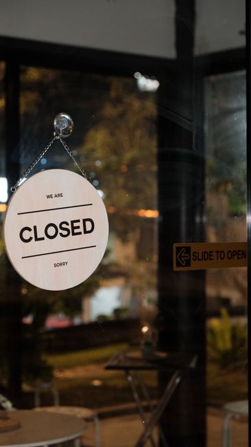 A circular white 'Closed' sign hanging by a chain from the door frame of an indoor space, indicating that the premises are not open. Behind the sign, blurred outdoor scenery is visible through large g