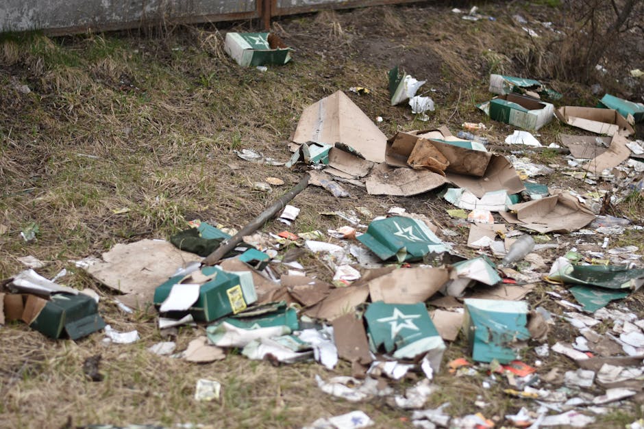 An outdoor scene showing a grassy area near a building with a large amount of scattered household waste and debris, including flattened cardboard boxes, torn packaging paper, and discarded cartons, some with visible branding. The waste is spread across the ground and partially extends onto a paved area, with a metal pole or rod lying among the rubbish. The background features a wall or fence, and the overall environment suggests illegal fly-tipping, often associated with improper disposal during house removals. The cluttered debris highlights issues related to improper waste disposal linked to home relocation activities, which Man with Van Coombe emphasizes in their removal services and disposal advice.