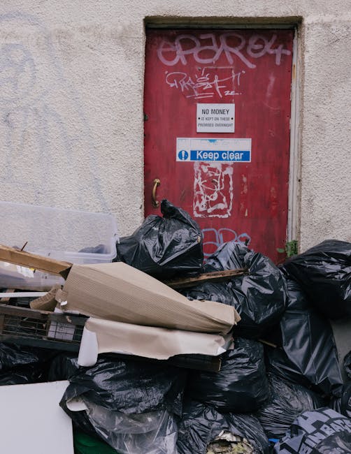 An outdoor area next to a building featuring a red metal door with graffiti and multiple notices, including 'No Money' and 'Keep Clear,' situated above a substantial pile of domestic waste. The waste includes black plastic rubbish bags, some torn open revealing contents, as well as flattened and unwrapped cardboard boxes, and packaging materials such as plastic wrap and foam. The waste is piled on the pavement directly in front of the door, obstructing access, and appears to be a result of illegal fly-tipping following a home relocation or furniture transport process. The building wall is plain with a textured surface, and lighting suggests daytime conditions. The presence of discarded packaging and waste outside the property highlights improper disposal, a risk often associated with move-related clearance or unprofessional disposal following a move. Man with Van Coombe, a provider of removals and house relocation services, emphasizes the importance of proper waste management during and after moving activities to avoid illegal dumping and potential penalties.