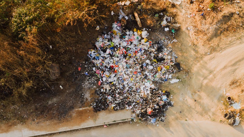 An outdoor scene showing a grassy area near a building with a large amount of scattered household waste and debris, including flattened cardboard boxes, torn packaging paper, and discarded cartons, some with visible branding. The waste is spread across the ground and partially extends onto a paved area, with a metal pole or rod lying among the rubbish. The background features a wall or fence, and the overall environment suggests illegal fly-tipping, often associated with improper disposal during house removals. The cluttered debris highlights issues related to improper waste disposal linked to home relocation activities, which Man with Van Coombe emphasizes in their removal services and disposal advice.