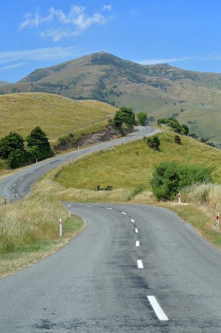 A rural winding road with two lanes, marked by dashed white lines, extends through a landscape of rolling green hills and scattered trees under a partly cloudy blue sky. In the foreground, the tarmac surface appears smooth, with roadside posts on each side. The road gently bends to the right and then to the left as it ascends into a hilly terrain. The scenery features grassy fields, bushes, and small patches of woodland, with a large hill or small mountain visible in the background. This setting depicts a peaceful countryside route suitable for vehicle travel, potentially used during a home relocation or furniture transport process. The image highlights the importance of choosing optimal local routes for efficient house removals, which [COMPANY_NAME] facilitates through careful planning of moving logistics and transport. The natural environment and road conditions reflect the scenic routes often used in house removals from Kingston Hill to Coombe, ensuring safe and smooth transportation of furniture and belongings.