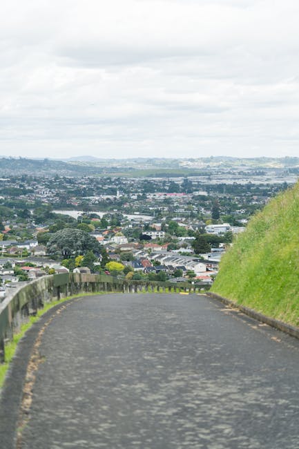 A paved, slightly curved road descending from a hillside, bordered by a metal guardrail on the left side and a green grassy slope on the right. The road appears to be used for home relocation or moving services, with no vehicles or people visible. In the background, there is a suburban landscape with numerous residential houses, trees, and commercial buildings, extending towards a distant horizon of rolling hills and a cloudy sky. The scene is well-lit with natural daylight, indicating daytime conditions. This setting demonstrates an accessible route for furniture transport, with the image representing the kind of local routes involved in house removals and moving logistics, as seen in services provided by Man with Van Coombe.
