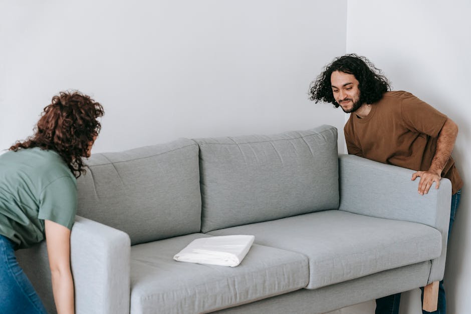 Two individuals are engaged in a home relocation process inside a room with plain white walls. The woman, with curly hair and wearing a green top, is bending forward, lifting or adjusting a light grey fabric-upholstered sofa. The man, with long curly hair and a beard, dressed in a brown t-shirt, is positioned on the right side of the sofa, holding the armrest to assist the movement. On the sofa's surface, there is a neatly folded white blanket or cushion. The scene appears well-lit with natural or soft indoor lighting, and the flooring is not visible in the image. This loading and moving activity reflects a furniture transport scenario typical during packing and moving procedures, which is often managed by professional services like Man with Van Coombe, specializing in house removals and furniture transport on narrow lanes such as Coombe's. The furniture is likely being prepared for transport, indicative of the logistics involved in home relocation.