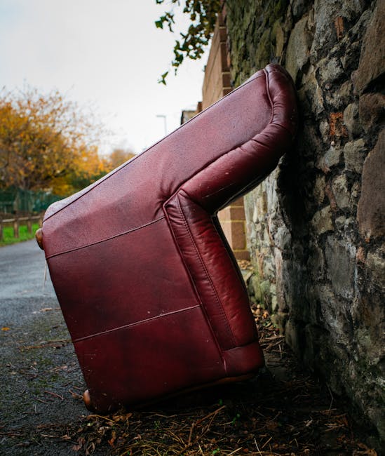A large, burgundy leather armchair has been placed upright against a stone wall on a narrow outdoor pavement in Coombe. The chair's backrest is angled towards the wall, with its seat and armrests visible. The surrounding environment includes a brick building on one side and rural landscape with trees in the background, indicating a residential area during autumn. The pavement appears to be part of a home relocation process, possibly awaiting loading onto a van, with no other furniture or packing materials visible in the scene. The natural lighting highlights the texture of the leather and the uneven surface of the stone wall, capturing a moment during furniture transport or moving preparations, which aligns with professional removals services offered by Man with Van Coombe.