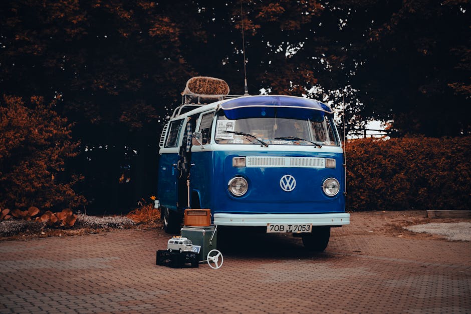 A vintage blue Volkswagen camper van parked outdoors on a paved surface with a background of dark foliage and trees. The van has a round Volkswagen emblem on the front and a rear-mounted spare tire with a brown cover. Items associated with home relocation are positioned near the front of the van, including a small black toolbox, a suitcase, and a toy model of a white van with a cargo container. The van's interior is partially visible through the open side door, indicating a setup for moving or packing activities. The scene is illuminated by natural daylight, highlighting the details of the vehicle and surrounding objects, reflecting a typical moving day setting involving furniture transport and packing preparations emphasized by [COMPANY_NAME], Man with Van Coombe, within the context of house removals and logistics.