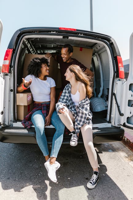 Inside the open rear of a large moving van parked outdoors, two women sit on the edge of the vehicle's loading area, engaged in conversation and smiling. One woman has curly black hair and is wearing a light blue t-shirt and jeans, gesturing with her hand. The other woman has long, light brown hair, and is dressed in a white top with a black and white plaid shirt tied around her waist, wearing light-colored tights and black-and-white sneakers. Behind them, a man stands inside the van, smiling as he interacts with the women. The van’s interior shows cardboard boxes and packing materials, including plastic wrapping and blankets, indicating a home relocation or furniture transport process. The scene is lit by natural daylight, and the setting suggests that the vehicle is prepared for a professional moving service. Man with Van Coombe is involved in packing and loading tasks, supporting efficient house removals with appropriate vehicle and equipment.