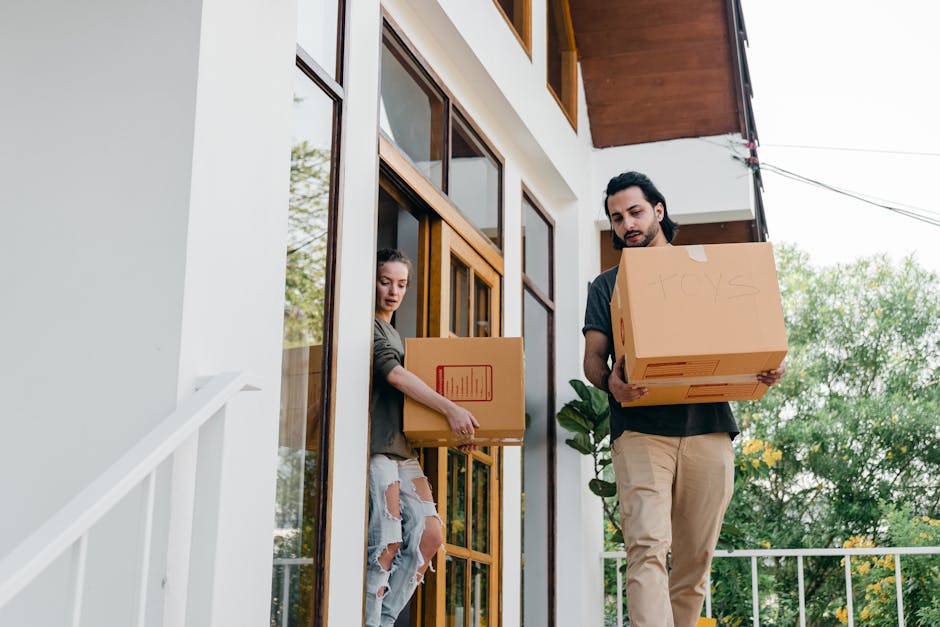 A man and a woman are carrying cardboard boxes filled with household items outside a modern house with large wooden-framed windows. The woman, dressed in a casual grey jacket and ripped jeans, holds a box labeled for moving or storage, standing on a balcony or porch area. The man, wearing a dark shirt and beige trousers, is holding a larger box labeled 'TOYS,' and both are in the process of loading or unloading belongings. The scene is set outdoors, with the house's white exterior walls and a wooden overhang visible, as well as greenery and trees in the background. The lighting indicates daylight, and the environment suggests an organized home relocation, with the boxes prepared for transport. The image reflects the logistics involved in packing and moving items during a house removal handled by [COMPANY_NAME].