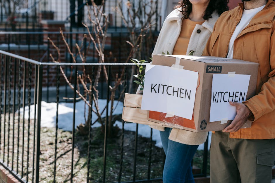 Two individuals are standing outside a residential property during daylight, each holding a cardboard box labeled 'KITCHEN' with a recycling symbol and a smaller wooden crate, indicating a home relocation or packing process. The person on the left, dressed in a cream sweater and blue jeans, is holding the wooden crate, while the person on the right, wearing an orange jacket and grey trousers, holds a large cardboard box with a label attached. The background features a black metal fence, leafless trees, and patches of snow on the ground, suggesting winter weather. The scene appears to be part of the loading or unloading stage of furniture transport and packing related to a moving service, with natural light illuminating the area. This image exemplifies aspects of professional removals and moving logistics, such as packing, handling, and preparing household items for transport, aligning with services offered by companies like Man with Van Coombe.