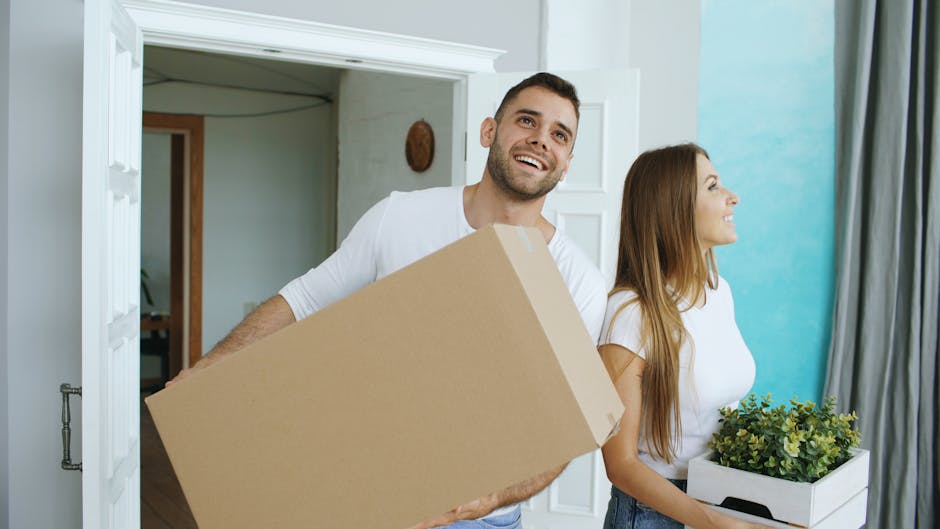 A man and a woman are carrying cardboard boxes filled with household items outside a modern house with large wooden-framed windows. The woman, dressed in a casual grey jacket and ripped jeans, holds a box labeled for moving or storage, standing on a balcony or porch area. The man, wearing a dark shirt and beige trousers, is holding a larger box labeled 'TOYS,' and both are in the process of loading or unloading belongings. The scene is set outdoors, with the house's white exterior walls and a wooden overhang visible, as well as greenery and trees in the background. The lighting indicates daylight, and the environment suggests an organized home relocation, with the boxes prepared for transport. The image reflects the logistics involved in packing and moving items during a house removal handled by [COMPANY_NAME].
